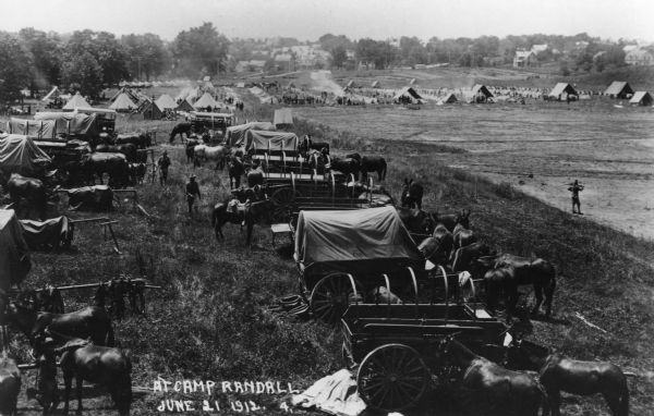 Slightly elevated view of Military training at Camp Randall. A number of horses and wagons line the foreground and rows of tents can be seen in the background. Caption reads: "At Camp Randall, June 21 1912. 4."
