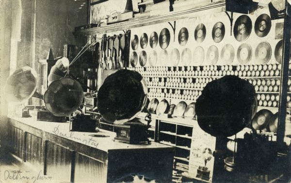 Interior view of a music shop with phonographs, violins, phonograph records displayed on the wall, and wax cylinders lining a shelf.