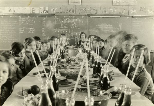 Students and teacher seated at a long table loaded with food and drinks for a Thanksgiving meal. There are music and English lessons behind them on the chalkboard.
