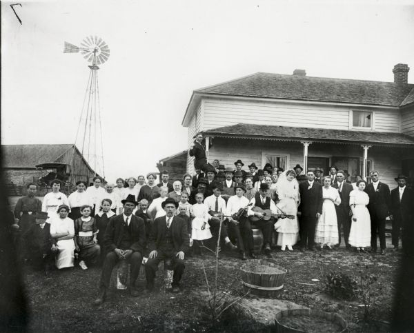 Family and friends gathered in front of a home for the wedding of Joseph Lorbieki and Agnes Onernek. Three musicians can be seen near the center. A windmill and a carriage can be seen in the background.