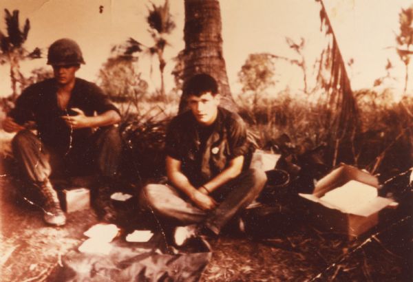 Soldier, possibly Michael Baldwin, sitting cross-legged at the base of a tree. Another soldier wearing a helmet is at the left.