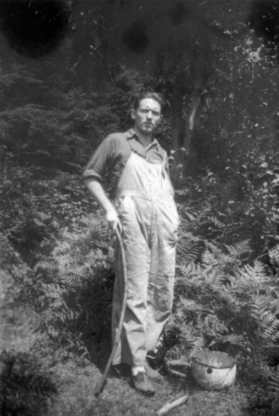 A casual portrait of Spencer Tracy wearing overalls and holding a long stick. There is a pot at his feet.