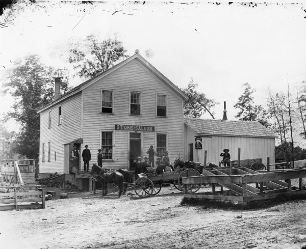 Exterior view from across road of men gathered outside a store and saloon. Most of the men hold a glass of beer as though toasting. Two teams of horses are hitched to wagons in front of the establishment. There is an advertisement for Jung and Borchert Lager Beer on the side of the building.