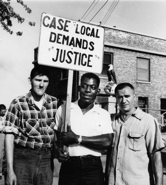 Three men pose together on a picket line at the Case plant. The man in the center holds a sign reading: "Case Local demands justice." The Case eagle on a globe insignia can be seen in the background. The men are members of UAW Local 180.