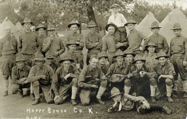 Group of soldiers of Co. K, 127th Infantry, 32nd Division, Wisconsin National Guard "at ease." Two men are eating, one holds a tin cup, two men hold mandolins, a man in back holds a young boy on his shoulder, and another boy lays on the ground at the front of the group. Tents can be seen in the background.