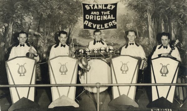 Portrait of Stanley and the Original Revelers, a five-piece band. The men are all dressed in white coats with dark collars and bow ties. Four of the musicians are seated behind podiums with the percussionist in the center. The backdrop is a woods scene.