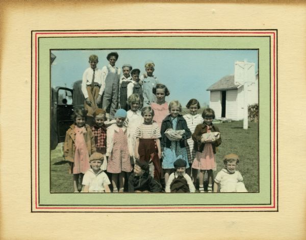Hand-colored photograph of students from Liberty school, a one-room school in Sheboygan County. The children in the back row are standing on the bumper of a car. A basketball hoop and shed are in the background.