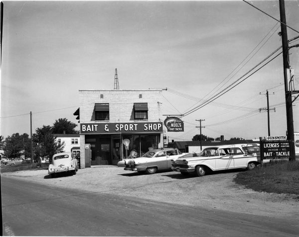 Exterior view of Noel's Bait and Sport Shop with cars parked in the lot in front of the building. Fishing nets can be seen on display in the window. The business has a neon sign with a trout on it.