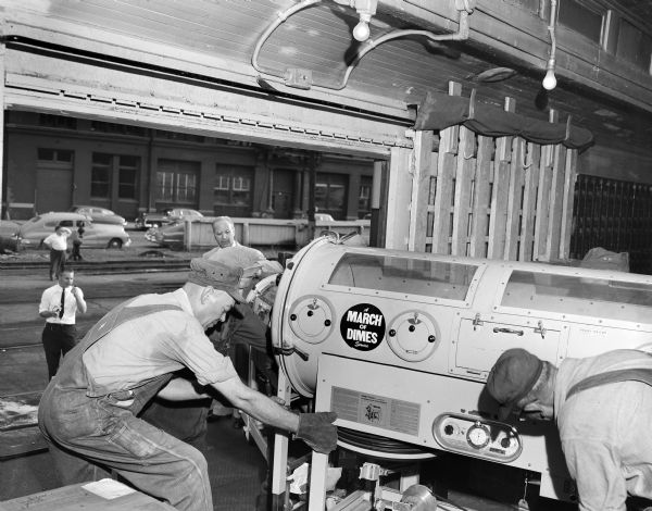 Men moving an iron lung. The machine has the logo for the March of Dimes imprinted on it. Iron Lungs were commonly used for the treatment of polio.