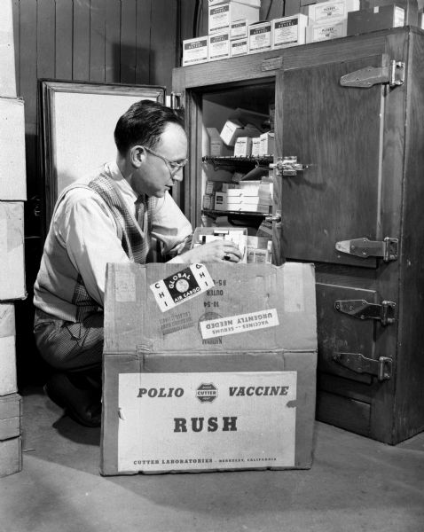 A doctor unloading a box filled with a supply of the polio vaccine. The box in the foreground is labeled: "Polio Vaccine Rush" and the doctor is kneeling near a refrigerated case used to store medication.