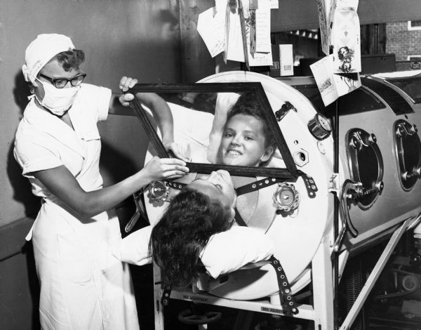A nurse holding a mirror so that the photographer can see the face of a girl in an iron lung. Hanging from the machine are a number of greeting cards. The caption on the back of the photograph reads: "Stricken with polio (she neglected to get her Salk shots) Flossie Rogers still managed a smile as she looked at the world through the mirror of her iron lung in June, 1957. So thankful for the care she got from the nurses at County Hospital in Mobile, Ala., Flossie vowed to devote her life to helping the sick if she ever got back on her feet. Helped with funds from the March of Dimes, Flossie did recover, and today is working to become a nurse at the same hospital."