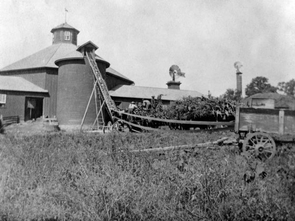 The barn in the foreground, left, is the school barn for the Hillside Home School (which was run by Frank Lloyd Wright's aunts & Enos Lloyd Jones sisters, Jennie & Nell Lloyd Jones). The windmill tower in the background, right, is Frank Lloyd Wright's 1896 design, the Romeo & Juliet Windmill Tower.  Several farm workers load corn from a wagon into a silo, which is in front of a barn. Behind the wagon is belt-driven machinery, which is powering the loader to lift the corn into the silo.