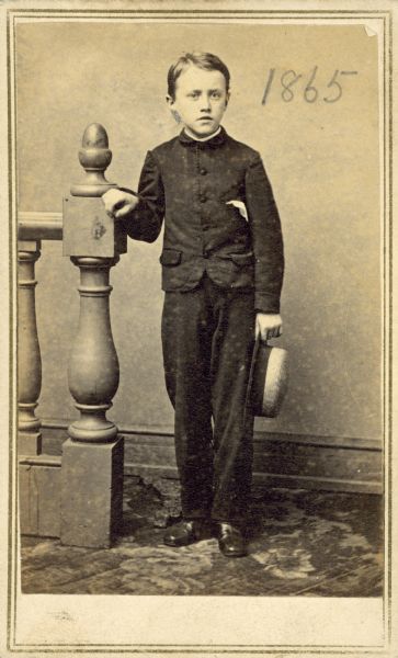 Full-length studio portrait of young George Hawthorne Scidmore standing with his hand on a newel post, and holding a hat in his other hand.