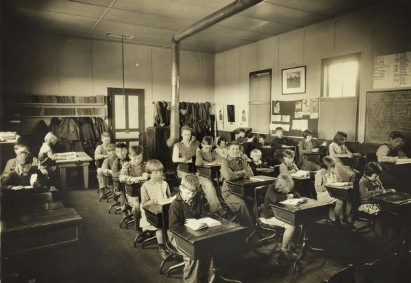 Interior view of Eagle Corners School, a one-room schoolhouse in Richland County. The students are all seated at desks and most are reading. Their coats hang on hooks at the back of the room near the stove.