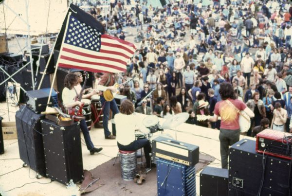 Wilderness Road during their performance at Sound Storm taken from the scaffolding behind the stage. Guitarist at right is Nate Herman, drummer is Tom Haban, bassist is Andy Haban, guitarist obscured by the U.S. flag is Warren Leming. An unidentified conga player can be seen between Leming and Haban. A portion of the crowd can be seen at the foot of the stage.