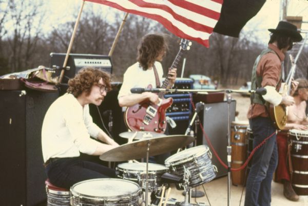 Wilderness Road taken from the stage during their performance at Sound Storm. The drummer is Tom Haban, bassist is Andy Haban, and the guitarist is Warren Leming. An unidentified conga player is sitting in with the band in the background. A U.S. flag and a black flag fly behind the band.