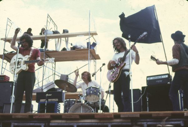 Wilderness Road seen from the foot of the stage during their performance at Sound Storm. The guitarist at left is Nate Herman, the drummer is Tom Haban, the bass player is Andy Haban, and the guitarist at right is Warren Leming. A black flag flies behind the band and scaffolding is set up at the back of the stage.