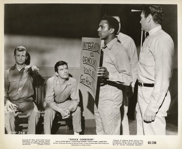 Movie still from the Allied Artists film "Shock Corridor," featuring James Best (playing Stuart), Peter Breck (playing Johnny Barrett), and Hari Rhodes (playing Trent). Trent, a black inmate of the psychiatric asylum, is standing in a hall with other patients. He carries a sign that reads, "Integration and Democracy don't mix. Go Home Nigger."