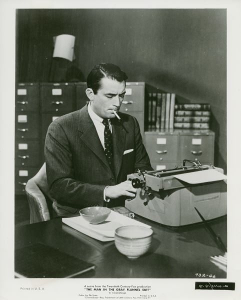 Movie still from the Twentieth Century-Fox movie "The Man in the Gray Flannel Suit," featuring Gregory Peck (playing Tom Rath). He is smoking a cigarette and using a typewriter in an office.