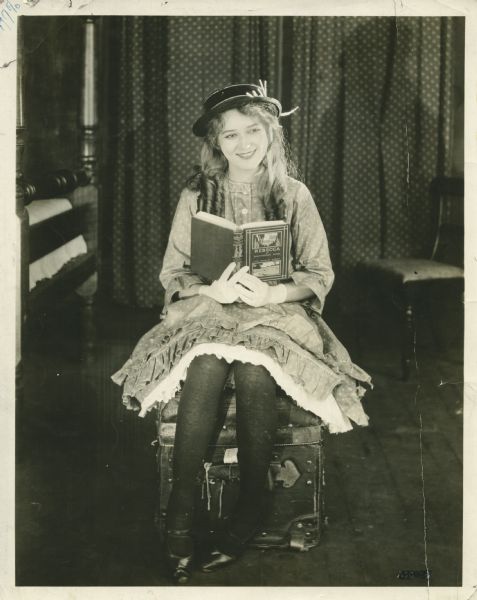 Publicity still for the Artcraft movie "Rebecca of Sunnybrook Farm," featuring Mary Pickford in the title role, in costume, reading the book by Kate Douglas Wiggin.