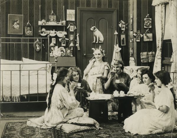 Scene still from the Lubin silent film "A Rebellious Blossom," featuring Florence Lawrence (playing the Rebellious Daughter) and other unidentified actresses enjoying tea in a dorm room decorated with many photographs, doodads, and Nipper, the dog trademark for Victor gramophone records.