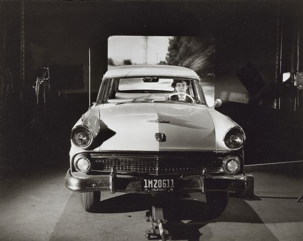 Production still showing the technique of rear projection. A 1951 Ford, jacked up in front so the steering wheel can turn, sits in front of a rear projection screen showing moving street scenes. When photographed from the front, the unidentified actor behind the wheel will appear to be driving a moving car instead of sitting still in the studio.