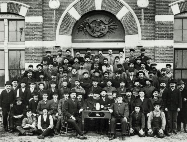 A large group of Brewery workers posing outside of Gund's Brewery in La Crosse. In the front of the group two men are seated at a table with a large stein and a few smaller glasses of beer.