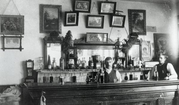 Saloon interior of two bartenders standing behind the bar. On the bar is a variety of glassware and bottles. On the wall behind the bar are three large mirrors along with a variety of framed prints of landscapes.