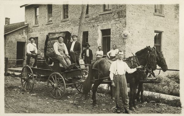 A group of men standing with a horse and wagon. Shown (L-R) Otto Petersilie, unknown, unknown, unknown, Joseph F. Faust, unknown, Charles J. Faust. The wagon is carrying a barrel of beer. The sign on the building in the background is an advertisement for beer. 