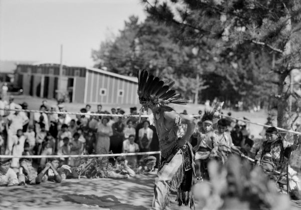 View of a Native American man dancing and wearing traditional dress and a headdress. A group of younger boys, some wearing traditional dress, watch him from the ropes lining the edge of the stage. A crowd of children and adults are watching in the background. In the far background are a building and trees.