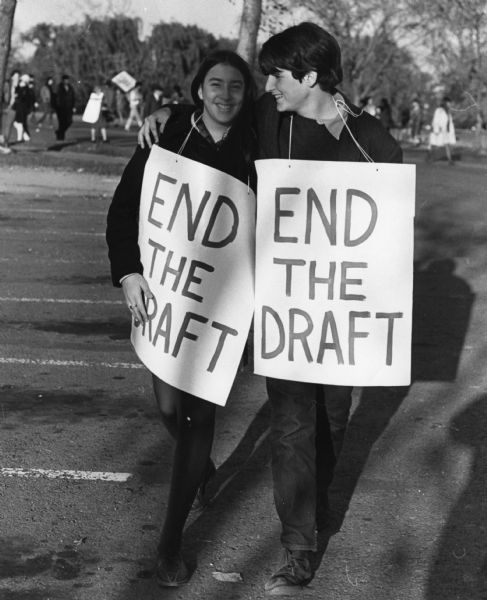 Couple walking towards the camera across a parking lot. They are wearing "END THE DRAFT" signs which are tied with string around their necks. He has his arm around her shoulders and they are smiling. Other demonstrators are visible in the background.