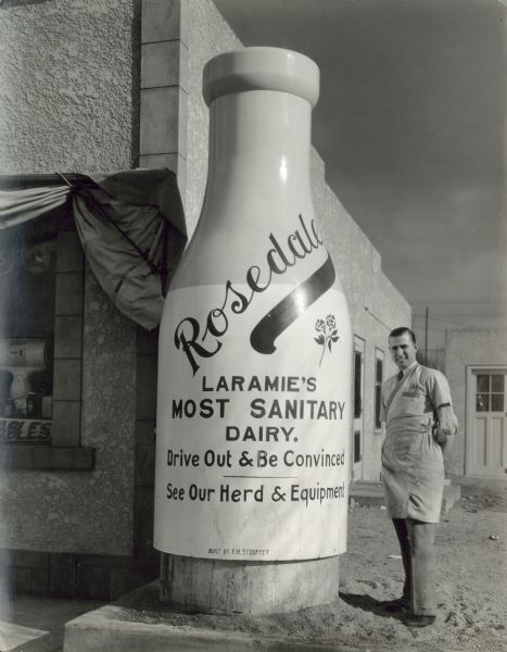 A smiling man stands outdoors next to a giant milk bottle upon which is painted "Rosedale, Laramie's Most Sanitary Dairy. Drive Out & Be Convinced. See Our Herd & Equipment".