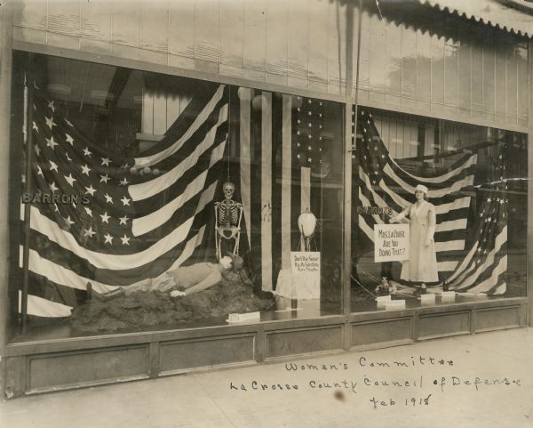 A window display by the Woman's Committee of the LaCrosse County Council of Defense showing a nurse, an injured soldier, a skeleton and American flags. The display is in the window of Doerflinger's Department Store on the corner of Main and 4th Streets.