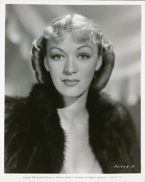 Publicity still of actress Eve Arden, wearing a fur coat. Caption reads: Different - is the word for this smart hairdress worn by Eve Arden, in Paramount's "Cocoanut Grove," starring Fred MacMurray and Harriet Hilliard. The coiffure was arranged by LaVaughn Speer, Paramount hairdresser and proves how becoming a page-boy roll may be when combined with a center part and a few softly curled bangs over the forehead.