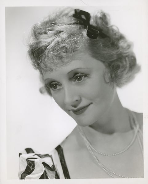 Publicity still of actress Billie Burke, with a black bow in her hair. Caption reads: The charm that is Billie Burke continues to captivate screen audiences the world over. The Metro-Goldwyn-Mayer actress is scheduled for many important roles in forthcoming pictures.