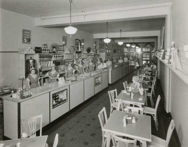 The interior of an ice cream shop, possibly Farmdale ice Cream Shop. Employees pose behind the long counters in uniform and several customers are seated at tables.
