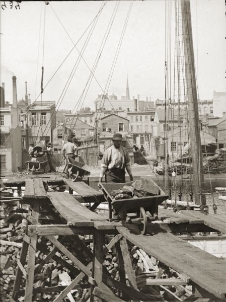 A man unloads coal from a boat by walking a wheelbarrow across wooden platforms raised on scaffolds. In the background a man stands with an empty wheelbarrow and two men fill a wheelbarrow with coal from a hanging barrel.