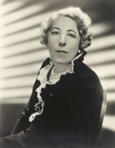Edna Ferber, Pulitzer Prize-winning novelist and playwright, in a publicity portrait. She is looking directly into the camera and is wearing a dress with a frilled neckline and a string of pearls with matching earrings.