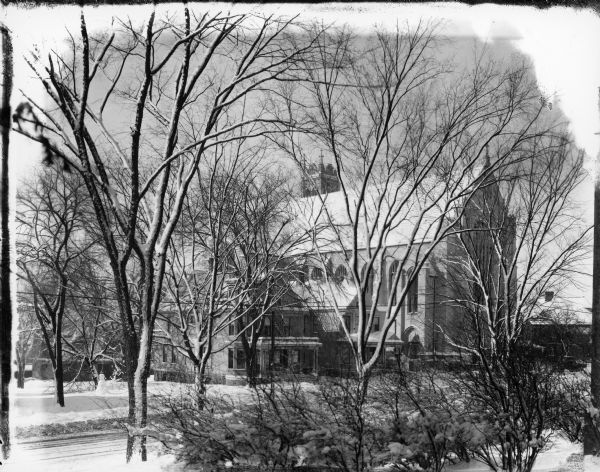 A view through snow-covered trees of Luther Memorial Church. There are two houses on the left side of the church, one has a sign above the porch that says "Saint Francis Club House." A snowman is in the yard.