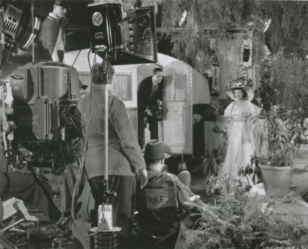 Scene still of a fictional camera and sound crew shooting Frederic March (playing actor Norman Maine) and Janet Gaynor (as Vicki Lester) on set in David O. Selznick's "A Star is Born" (Selznick 1937). March is emerging from a small travel trailer to join Gaynor who wears a long white dress and sun hat. This scene is a film within the film with Casey Burke (played by Owen Moore) sitting in the director's chair.