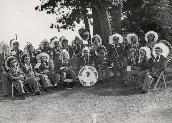 A group portrait of a band of Menominee Indians posing with their instruments. They are wearing feather headdresses.