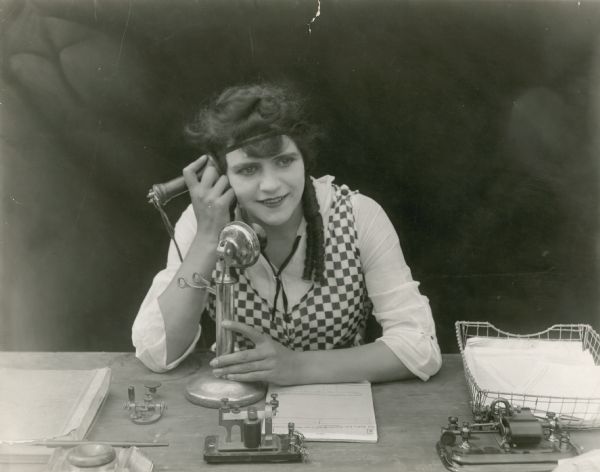Helen Gibson stars in one of the "Hazards of Helen" films. She sits at a desk holding the receiver of a candlestick telephone to her ear.