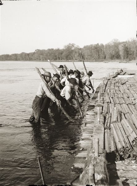 Stereograph of a group of men standing knee deep in water using handspikes probably in an attempt to dislodge a lumber raft from a sandbar or from the shoreline.