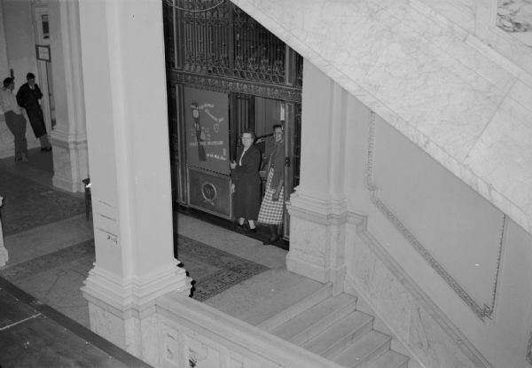 Elevated view from stairway of the ornate Otis elevator in the Wisconsin Historical Society building. This elevator was installed as original equipment in 1900. This view was taken of the first floor during remodeling, and it shows the elevator operator, Matilda Severson (left), and Willie Jo Walker, a member of the maintenance staff. A poster mounted on the front of the elevator says: "take a trip through Wisconsin's past VISIT THE MUSEUM on the 4th floor." Two men stand near a doorway in the background.