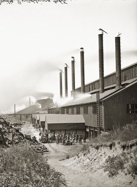 Exterior of the Bay View Rolling Mill, Puddling Department, with workers posing outside the building. The men are standing near railroad tracks, and a few of the men are sitting on a small railroad car.