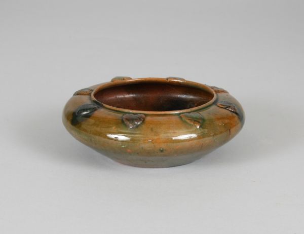 This earthenware bowl, which was designed by Susan Frackelton and thrown by George Ohr, is squat and bulbous. It is red with mottled green glaze on the exterior and clear and dark brown glazes on the interior. The bowl is decorated with molded hearts, diagonal bands, and flowers applied around the rim. A maker's mark, "SF," and date, "99," are incised on the underside, along with "G.E. Ohr" over "Biloxi, Miss" stamped in clay.

