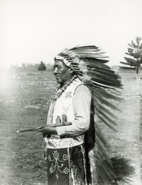 The image shows a performer standing and holding a pipe. Member of the Peavey Falls group of dancers and musicians from the Menominee Indian Reservation in Wisconsin.