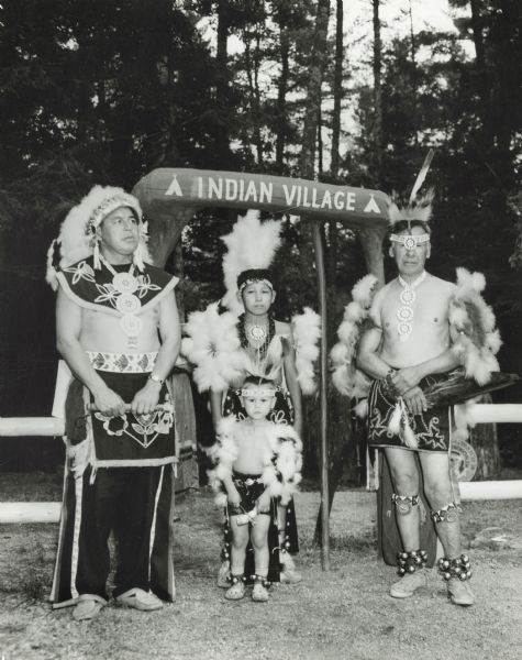 Four generations of men and boys pose for a  portrait of Menoninee Indians in full dress standing outdoors near a gate and fence. A sign says: "Indian Village". Members of the Peavey Falls group of dancers and musicians from the Menominee Indian Reservation in Wisconsin.
