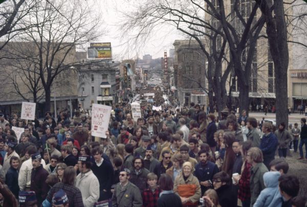 A large crowd participating in a demonstration against the war in Vietnam, as seen from the steps of the Wisconsin State Capitol looking down State Street toward the university. Many crowd members are wearing garrison style hats reading "Vietnam Vets For Peace." Businesses visible include Cupid's Corner, Rennebohm's, Commercial State Bank, Leath's, Wards, and the Orpheum Theatre.