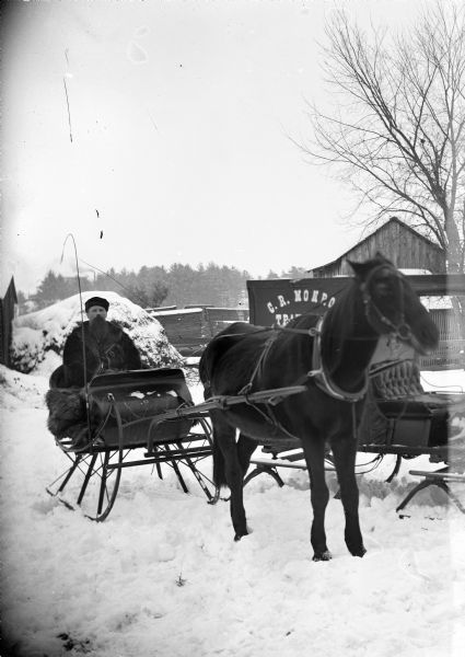 Winter scene with a European American man with a beard and moustache sitting in a sleigh pulled by a single horse on snow-covered ground in front of the C.R. Monroe Photowagon on runners. There is a barn in the background.